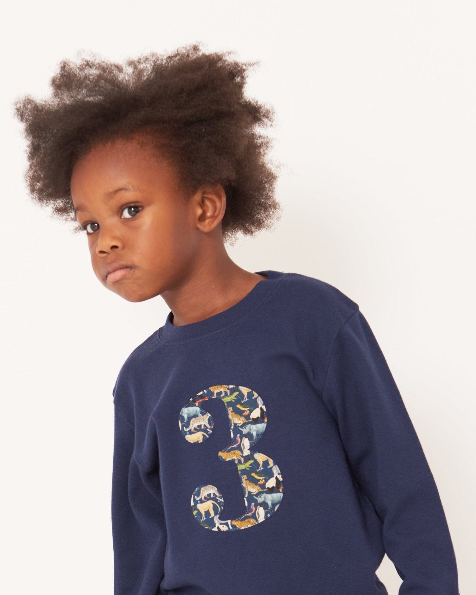 A 3 year old boy wearing a navy long sleeve t-shirt with a  number 3 on the front on in Liberty Quey Zoo animal print fabric.