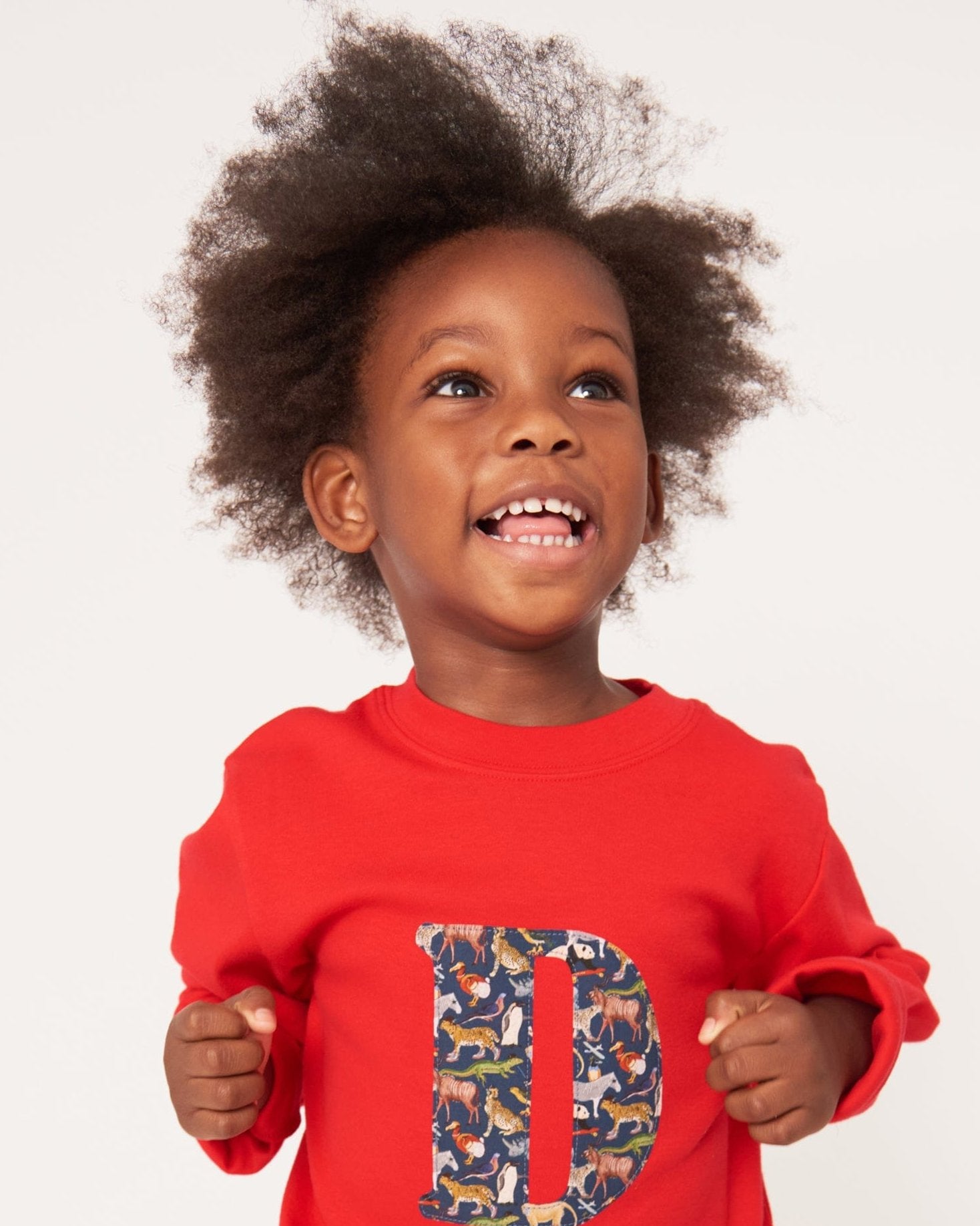 A smiling 2 year old boy wearing a personalised red t-shirt with his initial on the front made out of a Liberty zoo animal print Quey 2.