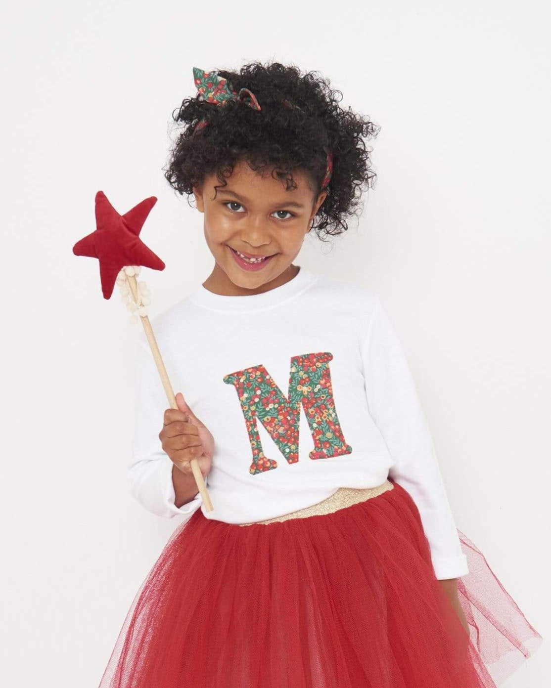 A 5 year old girl wearing a personalised white t-shirt with a her initial on the front made from Liberty Glitter Wiltshire berry fabric.