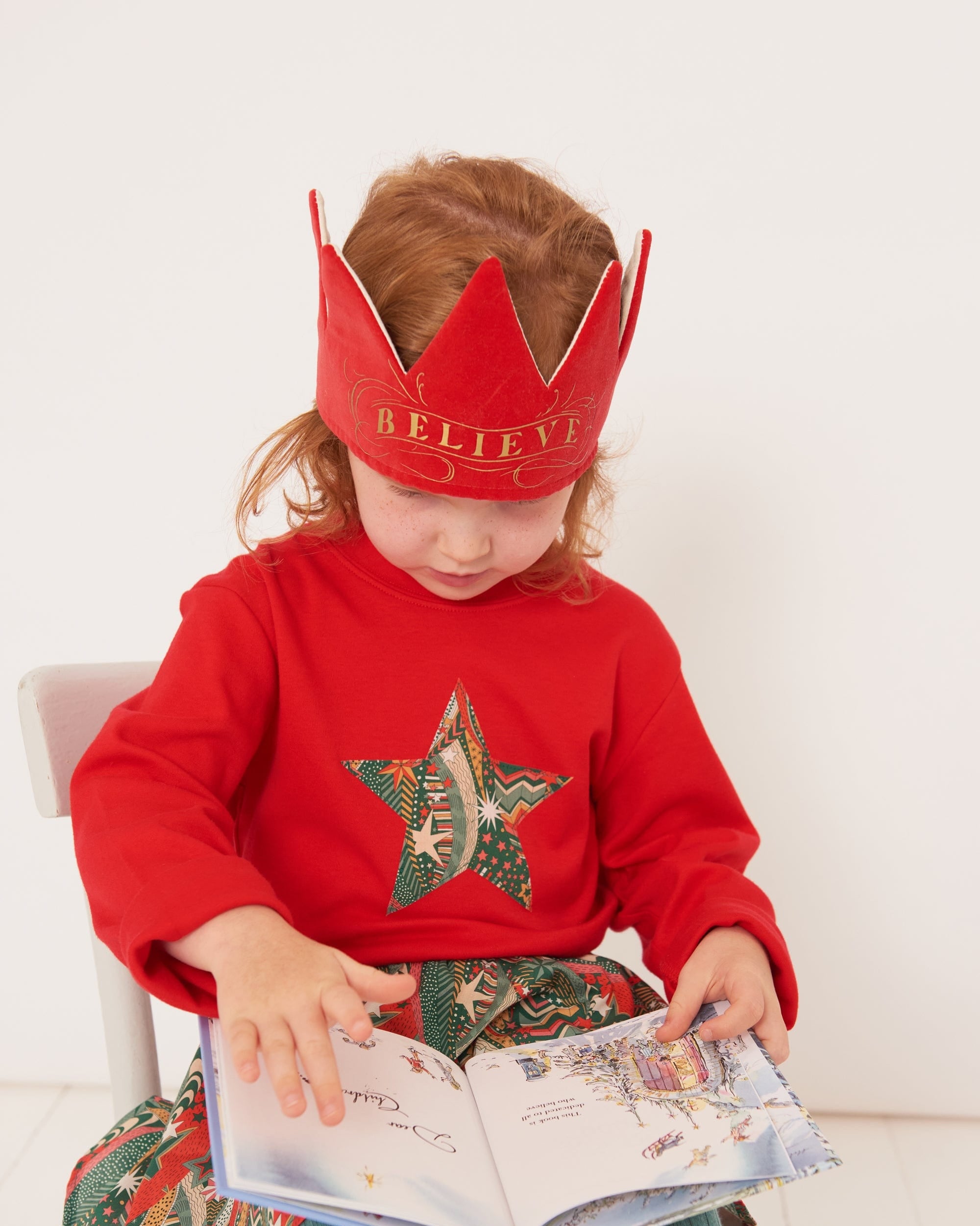 A 4 year old girl wearing a red Christmas t-shirt with a festive star on the front in a starry Liberty print, wearing a crown and reading a christmas book.