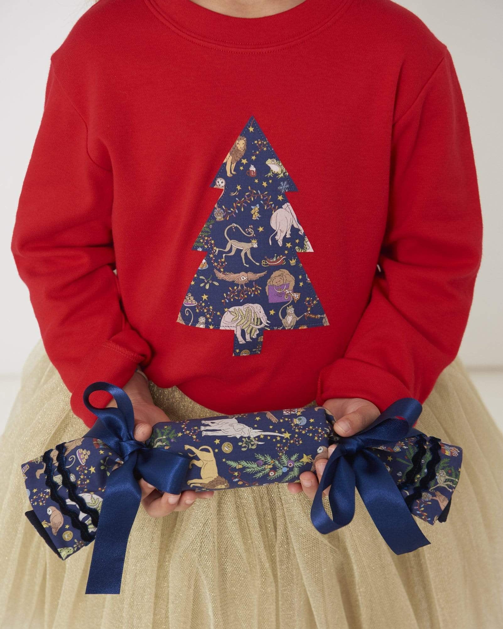 A girl holding a liberty cracker wearing a festive red t-shirt with a christmas tree on the front made from Liberty Christmas fabric.