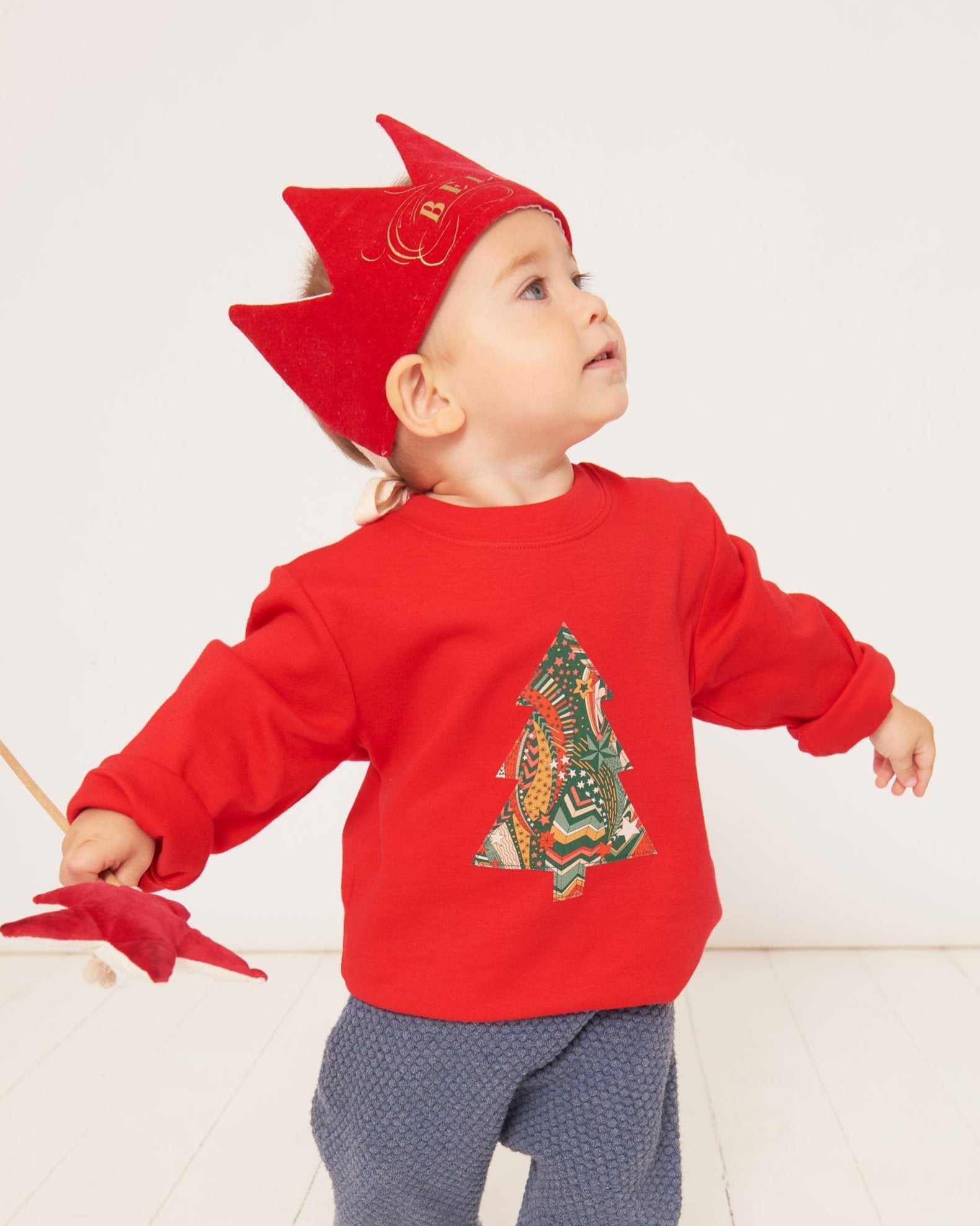 a one year old boy dancing in a festive red cotton t-shirt with a Christmas tree made from a stray liberty print on the front. He is holding a wand and wearing a red velvet crown.