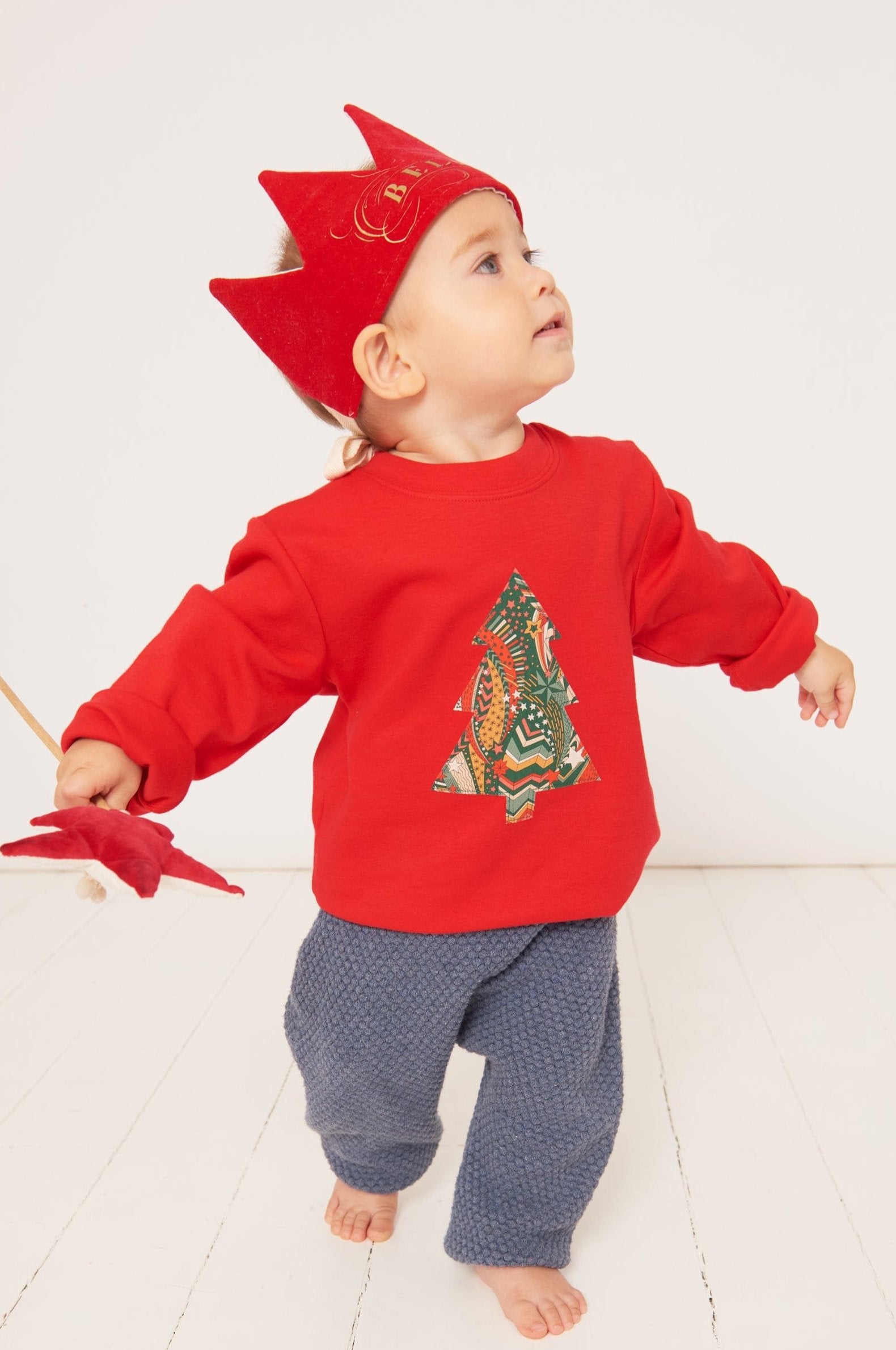 a one year old boy dancing in a festive red cotton t-shirt with a Christmas tree made from a stray liberty print on the front. He is holding a wand and wearing a red velvet crown.