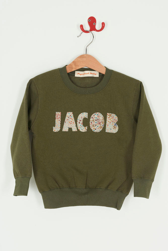 Green sweatshirt with 'JACOB' sewn on it in Liberty star Adelajda fabric, hanging on a wooden hanger against a white background.