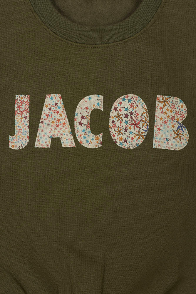 Green sweatshirt with 'JACOB' sewn on it in Liberty star Adelajda fabric, hanging on a wooden hanger against a white background.
