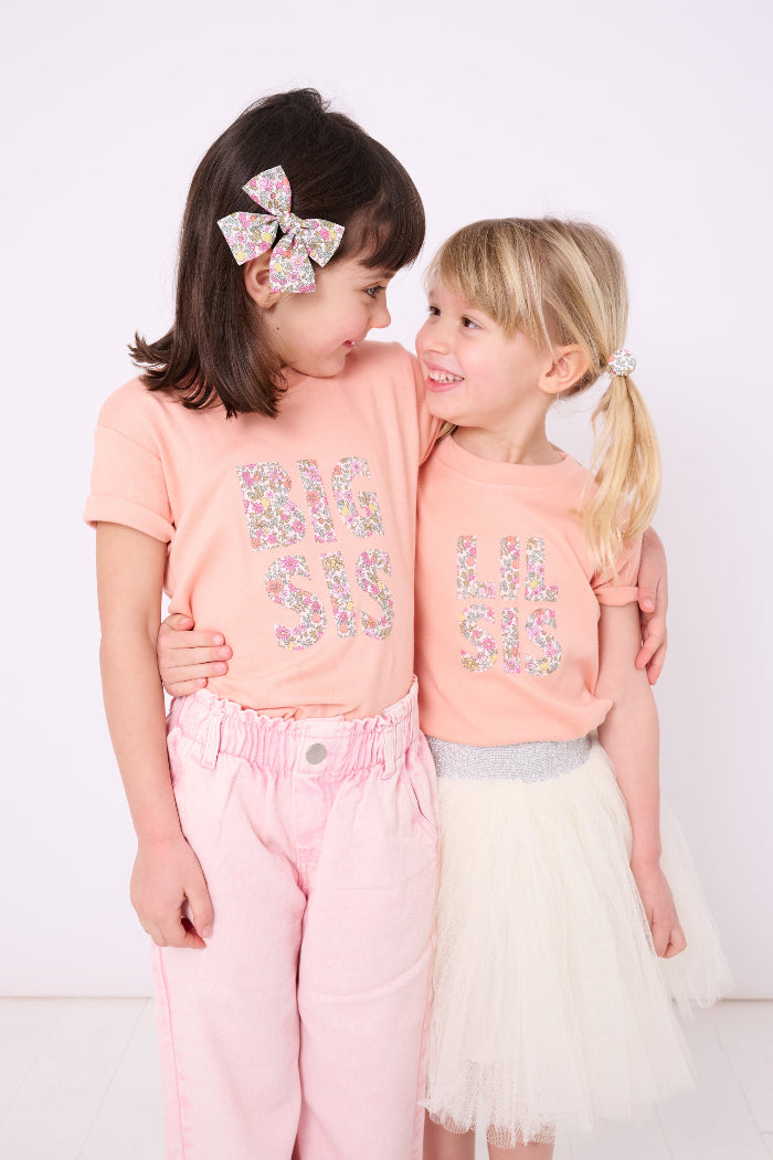 A girl wearing a BIG SIS pale Pink T-Shirt in Choice of Liberty Print as a proud big sister and a little sister wearing a 'lil sis' tee