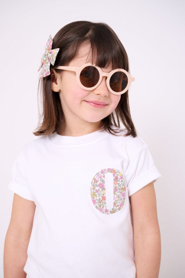 A Child wearing a white t-shirt with a small floral liberty print initial on the front.