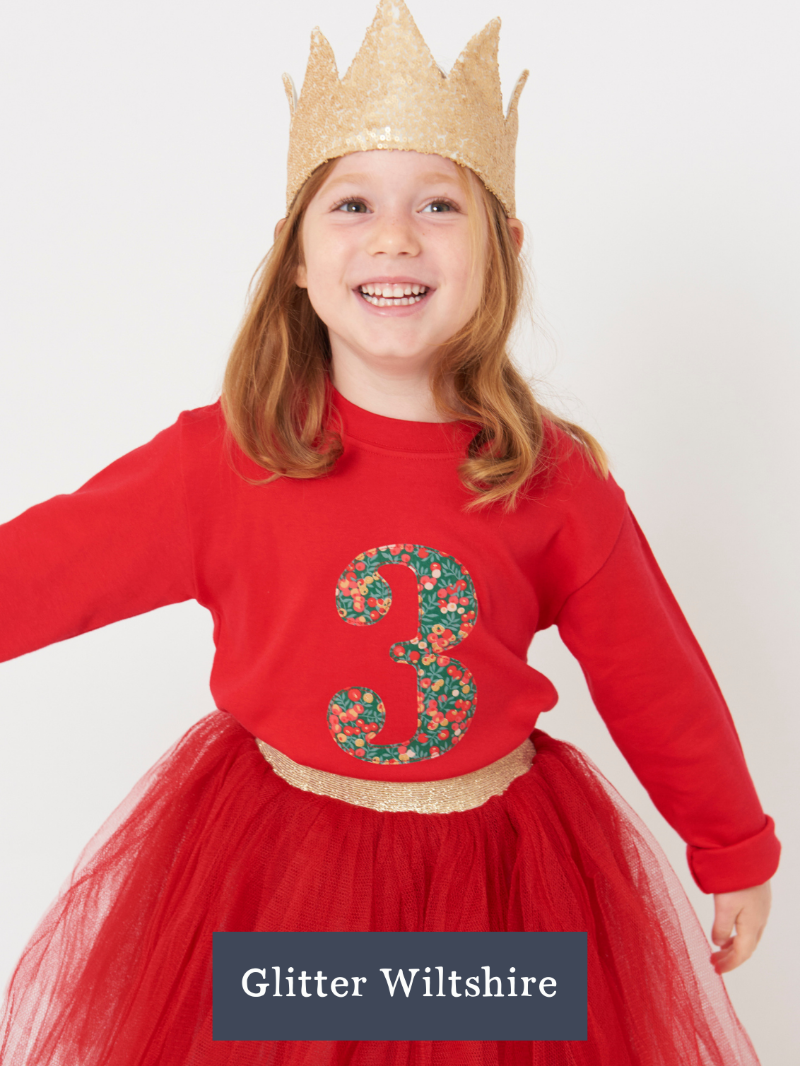 Child wearing a red t-shirt with the number 3 in liberty Wiltshire print, gold crown, and red skirt on a white background.