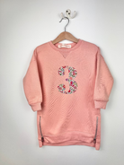 A pink sweatshirt hanging on a wooden hanger against a light gray wall. The sweatshirt features a large floral number 3 on the front and silver zipper details on the sides.