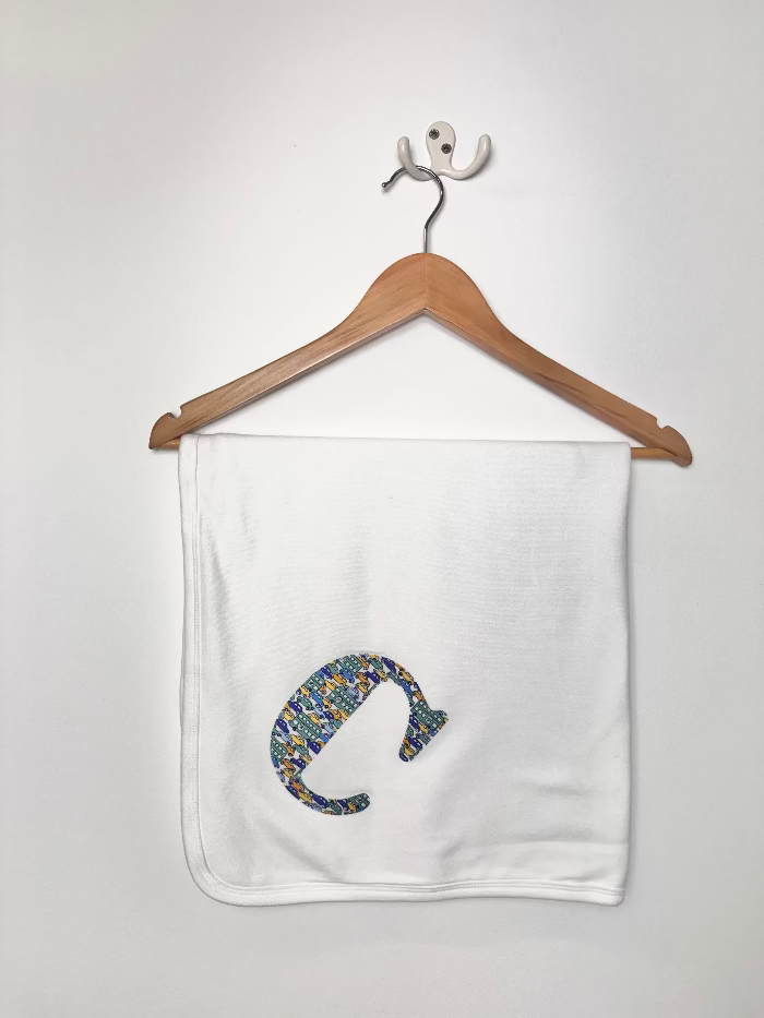 A white baby blanket hanging from a wooden hanger on a white wall. The blanket features a colorful, textured LIberty car print applique of the letter 'C' in blue, yellow, and green.