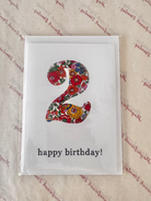 A square white birthday card in clear plastic packaging, featuring a large floral-patterned number 2 and the text 'happy birthday!' in black lowercase letters, placed on a beige patterned surface.