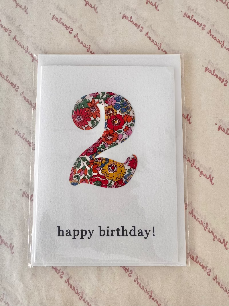 A square white birthday card in clear plastic packaging, featuring a large floral-patterned number 2 and the text 'happy birthday!' in black lowercase letters, placed on a beige patterned surface.