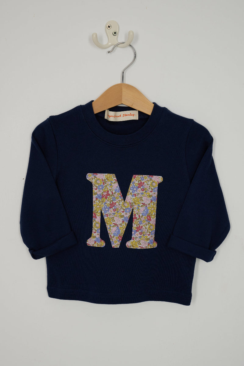 A navy blue long-sleeve t-shirt hanging on a wooden hanger with a white wall hook behind it. The sweatshirt features a large capital letter 'M' on the front filled with a Liberty print floral cotton