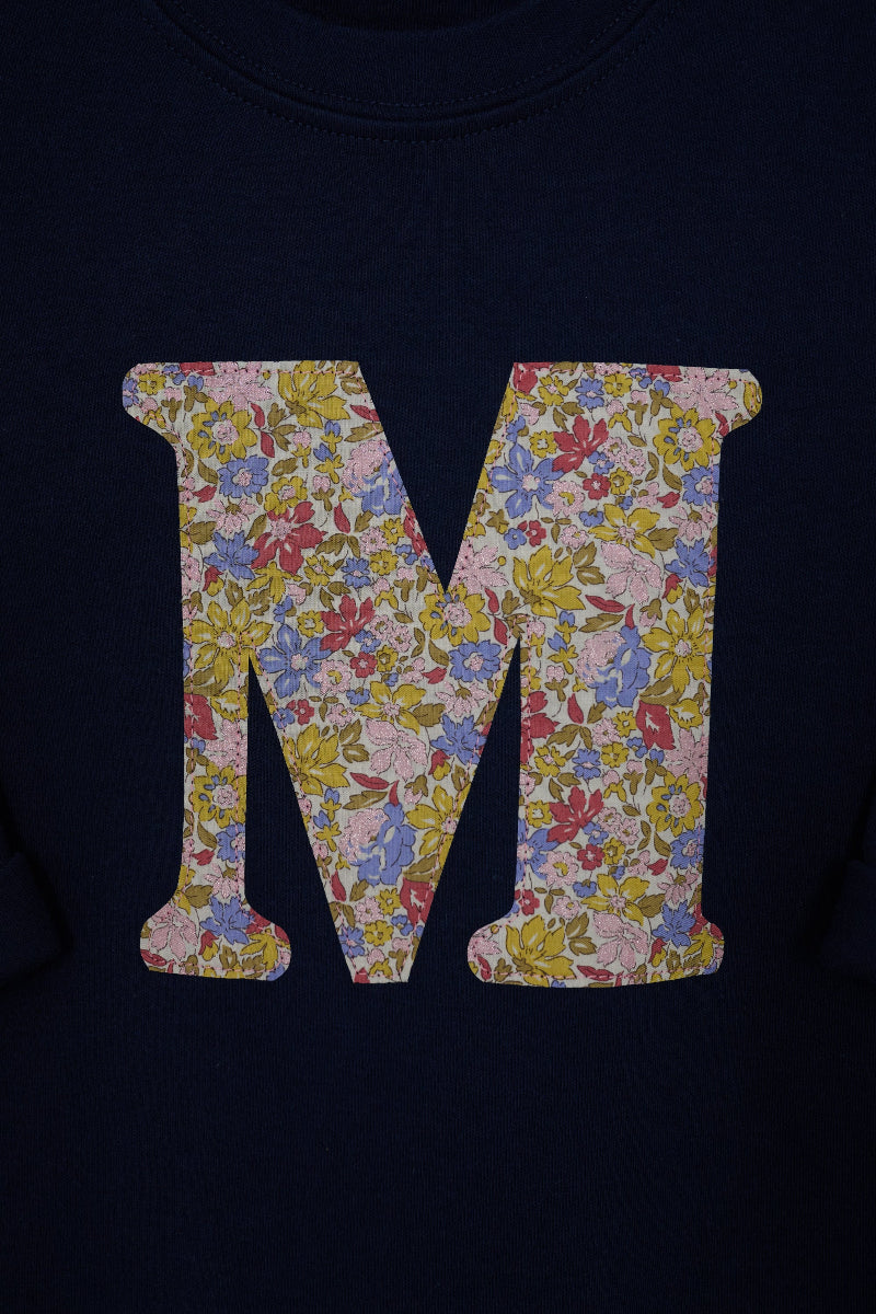 A close-up of a navy blue kids t-shirt featuring a large letter 'M' graphic filled with a Liberty print floral Juila Belle pattern.