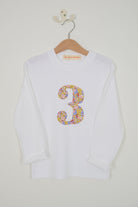 A white long-sleeve t-shirt with a large, colourful floral-patterned number 3 sewn on the chest cut from Liberty Julia Belle fabric, hanging on a wooden hanger against a plain off-white wall.