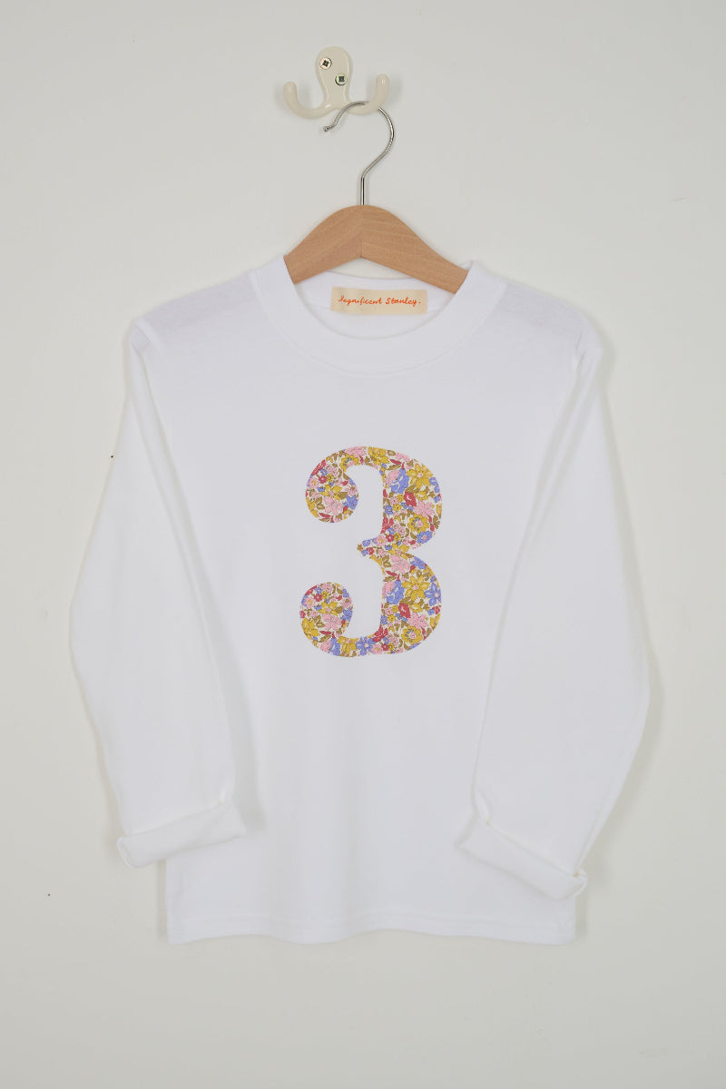 A white long-sleeve t-shirt with a large, colourful floral-patterned number 3 sewn on the chest cut from Liberty Julia Belle fabric, hanging on a wooden hanger against a plain off-white wall.