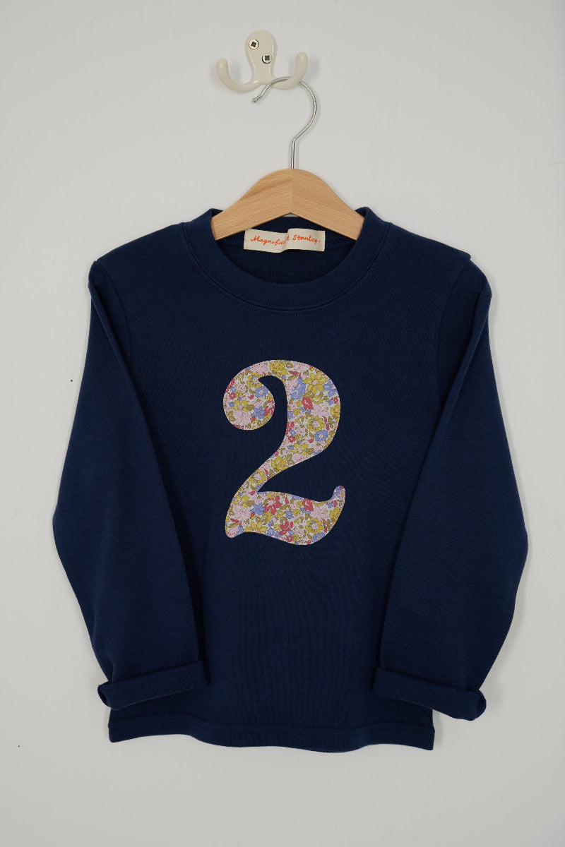 A navy blue long-sleeve sweatshirt hanging on a wooden hanger against a light gray wall. The t-shirt features a large, navy stylized number '2' on the front made from Liberty Julia Belle cotton floral pattern.