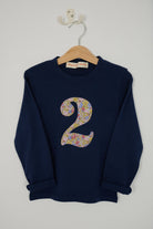 A navy blue long-sleeve sweatshirt hanging on a wooden hanger against a light gray wall. The t-shirt features a large, navy stylized number '2' on the front made from Liberty Julia Belle cotton floral pattern.
