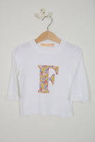 A white long-sleeve t-shirt with a large Juila Belle Liberty floral-patterned letter 'F' on the front, hanging on a wooden hanger against a plain off-white wall.