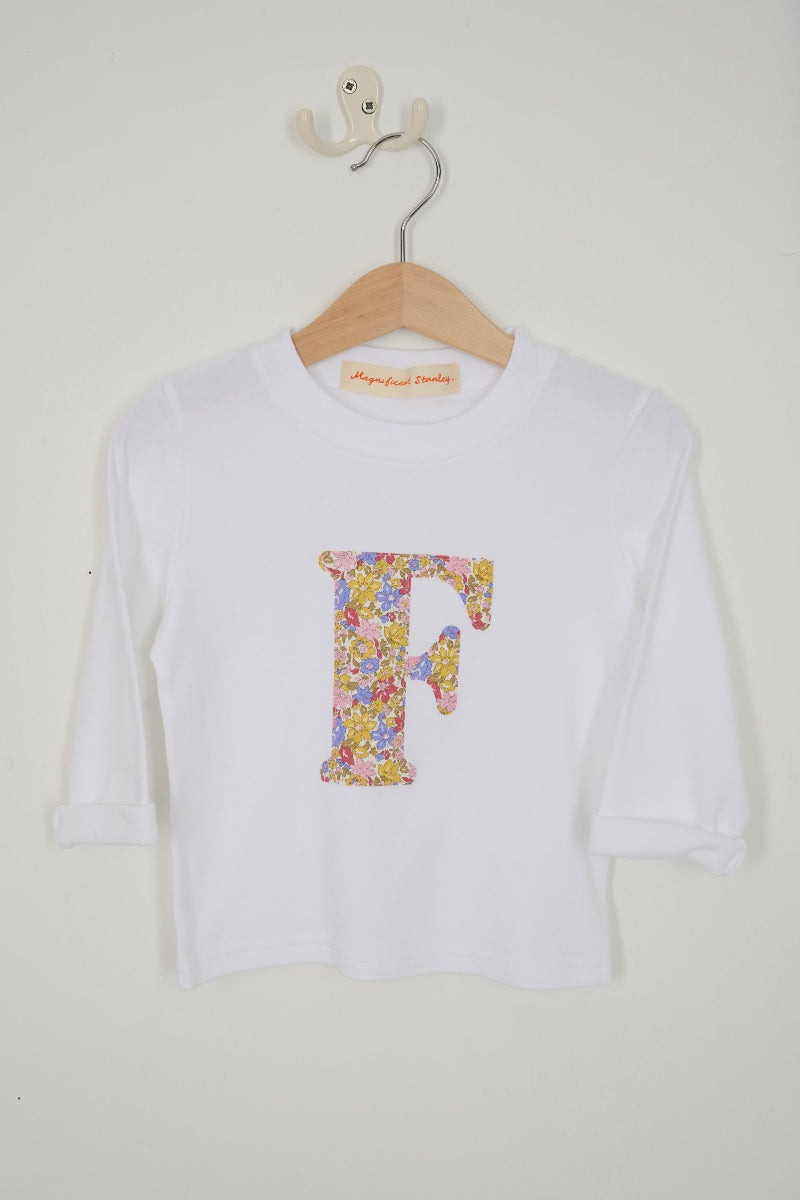 A white long-sleeve t-shirt with a large Juila Belle Liberty floral-patterned letter 'F' on the front, hanging on a wooden hanger against a plain off-white wall.