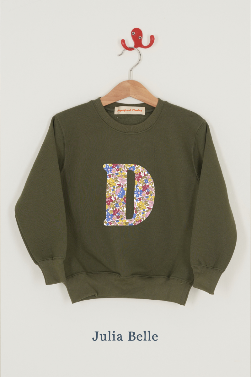 An olive green crewneck sweatshirt hanging on a wooden hanger with a red hook, featuring a large Liberty print floral-patterned letter 'D' on the front. The fabric name 'Julia Belle' is printed below the sweatshirt.