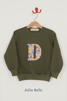 An olive green crewneck sweatshirt hanging on a wooden hanger with a red hook, featuring a large Liberty print floral-patterned letter 'D' on the front. The fabric name 'Julia Belle' is printed below the sweatshirt.