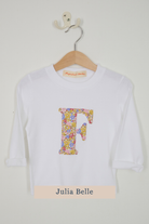 A white long-sleeve t-shirt with a large floral-patterned letter 'F' on the front, hanging on a wooden hanger against a light wall. A beige label at the bottom reads 'Julia Belle'.