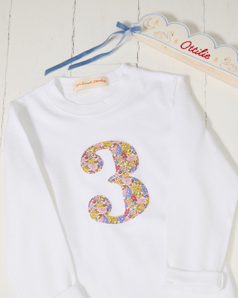 A white long-sleeve kids birthday t-shirt laid flat on a white wood background, featuring a large embroidered number 3 on the front filled with a floral Liberty pattern.