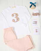 A children's outfit set laid out on a white wood background, including a white long-sleeve shirt with a large Liberty floral-patterned number 3 on the front, light pink jeans, a matching floral Liberty hair scrunchie, and matching hair clips and a meri meri party hat.