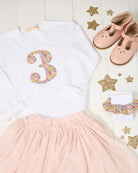 A styled flat-lay of a girl's birthday outfit set featuring a white long-sleeve top with a glittery pastel number 3, a light pink tulle skirt, a pair of pink Mary Jane shoes, a matching Liberty print pink scrunchie, and gold glitter star decorations on a white wood plank background.