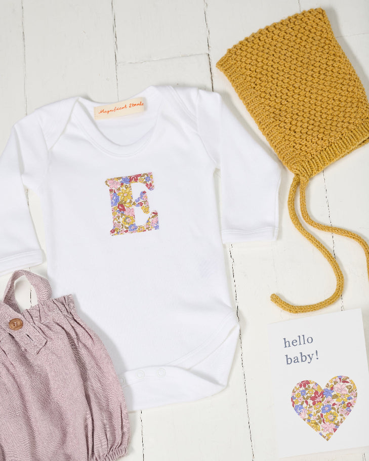 A baby outfit flat lay featuring a white long-sleeve bodysuit with a colorful floral Liberty letter 'E' on the chest, dusty pink overalls, a mustard yellow knit bonnet, and a 'hello baby!' card on a white wood plank background.