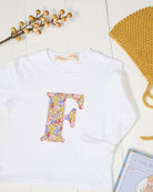A styled flat-lay featuring a white long-sleeve baby shirt with a colorful floral letter 'F' applique on the chest in LIberty Julia Belle print, accompanied by a mustard yellow knit bonnet, a pink hair tie with floral beads, and a children's book on a white wood plank background.