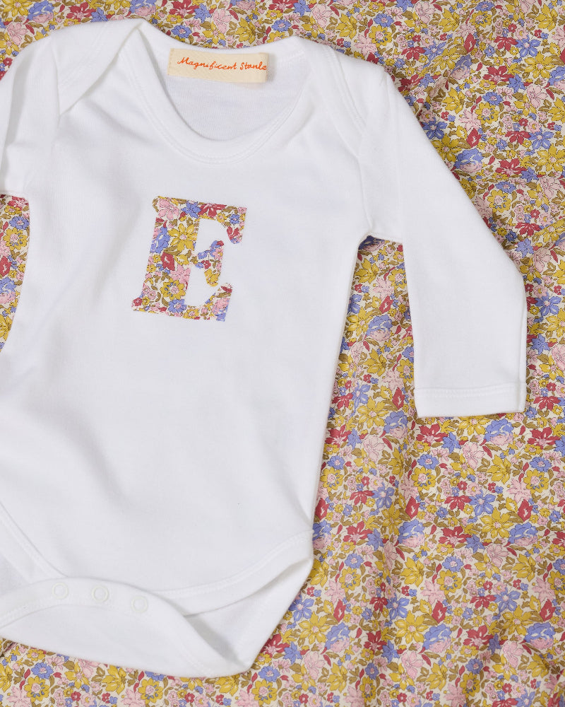 A white long-sleeve baby bodysuit laid flat on a yellow and pink sparkly Liberty Julia Belle floral fabric background. The bodysuit has a decorative floral block-letter 'E' on the chest.