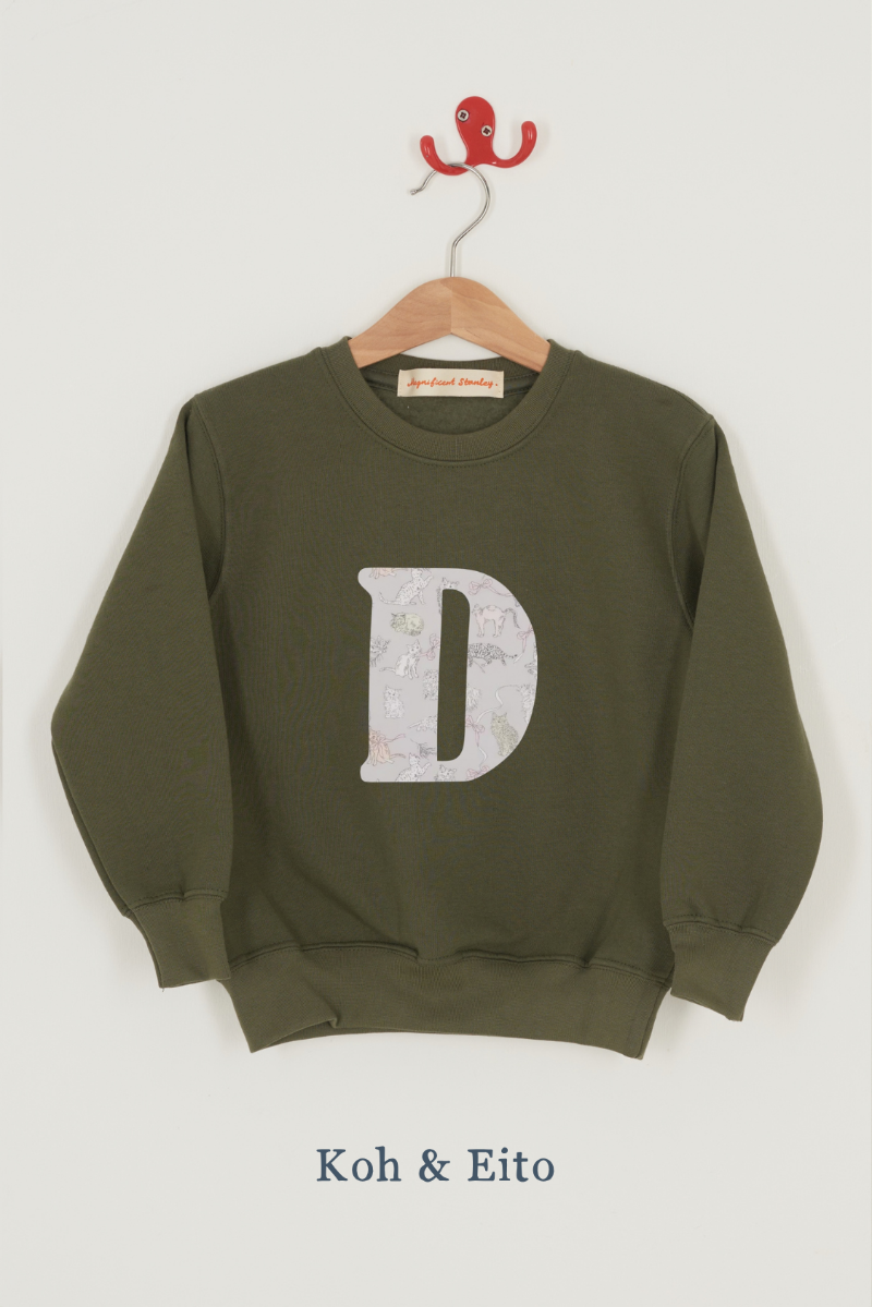 A khaki green crewneck sweatshirt hanging on a wooden hanger with a red hook, featuring a large Liberty print cat patterned letter 'D' on the front. The fabric name 'Koh & Eito' is printed below the sweatshirt.
