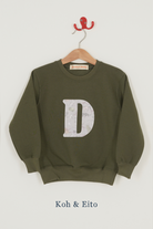 A khaki green crewneck sweatshirt hanging on a wooden hanger with a red hook, featuring a large Liberty print cat patterned letter 'D' on the front. The fabric name 'Koh & Eito' is printed below the sweatshirt.