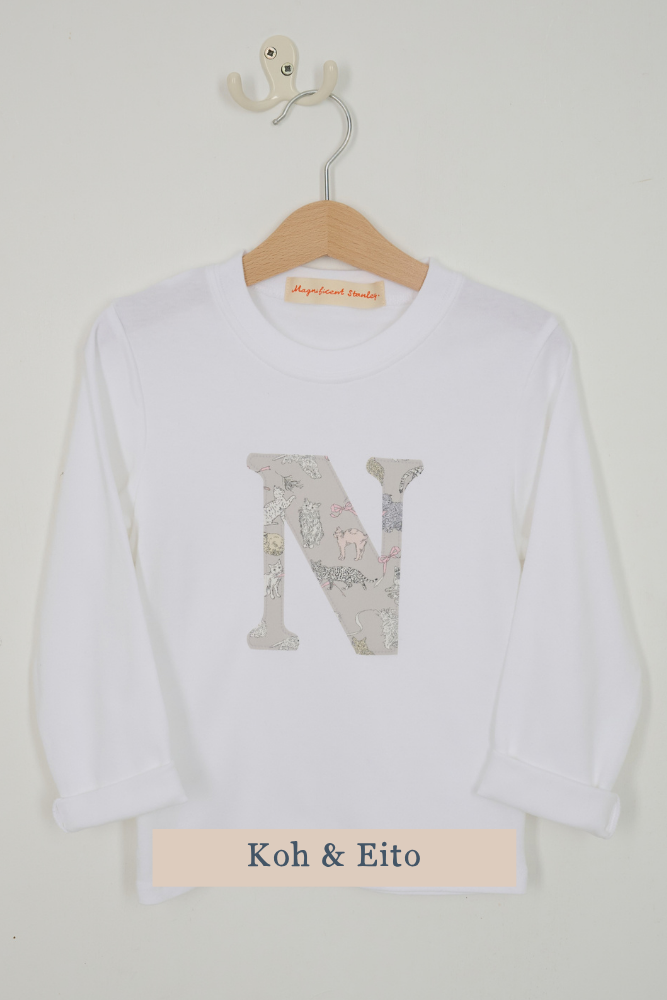 A white long-sleeve t-shirt with a large decorative letter 'N' on the front filled with a Liberty cats pattern, hanging on a wooden hanger against a light beige wall. A label at the bottom reads 'Koh & Eito'.