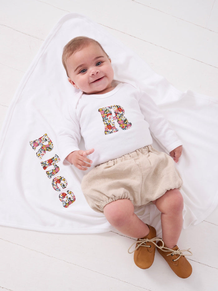 A newborn baby girl wearing a Personalised baby Bodysuit in Libby floral Liberty Print. She is laying on a personalised cotton baby blanket with her name sewn on.