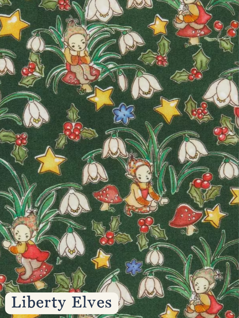 A repeating pattern of Liberty Elves fairies in red dresses among white snowdrops, red berries, green holly leaves, yellow stars, and red-and-white mushrooms on a dark green background. The text 'Liberty Elves' appears in a white rounded rectangle at the bottom left.