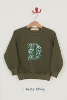 An olive green crewneck sweatshirt hanging on a wooden hanger . The sweatshirt features a large Liberty print Elves letter 'D' on the front filled with a colourful floral pattern featuring snowdrops, stars and holly.