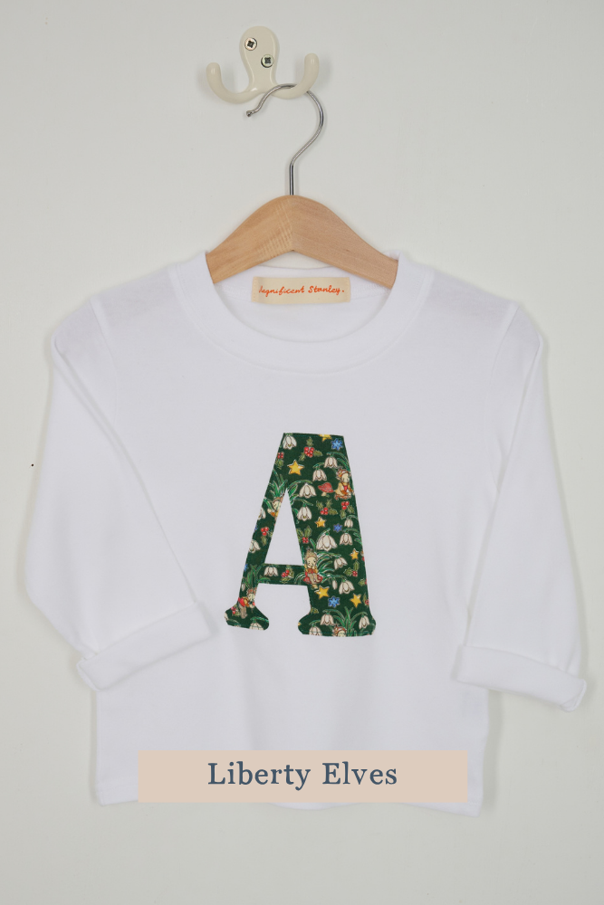 A white long-sleeve top for kids hanging on a wooden hanger against a light gray wall. The shirt features a large capital letter 'A' on the chest filled with a green floral Liberty Elves pattern.