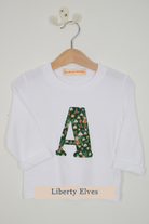 A white long-sleeve top for kids hanging on a wooden hanger against a light gray wall. The shirt features a large capital letter 'A' on the chest filled with a green floral Liberty Elves pattern.