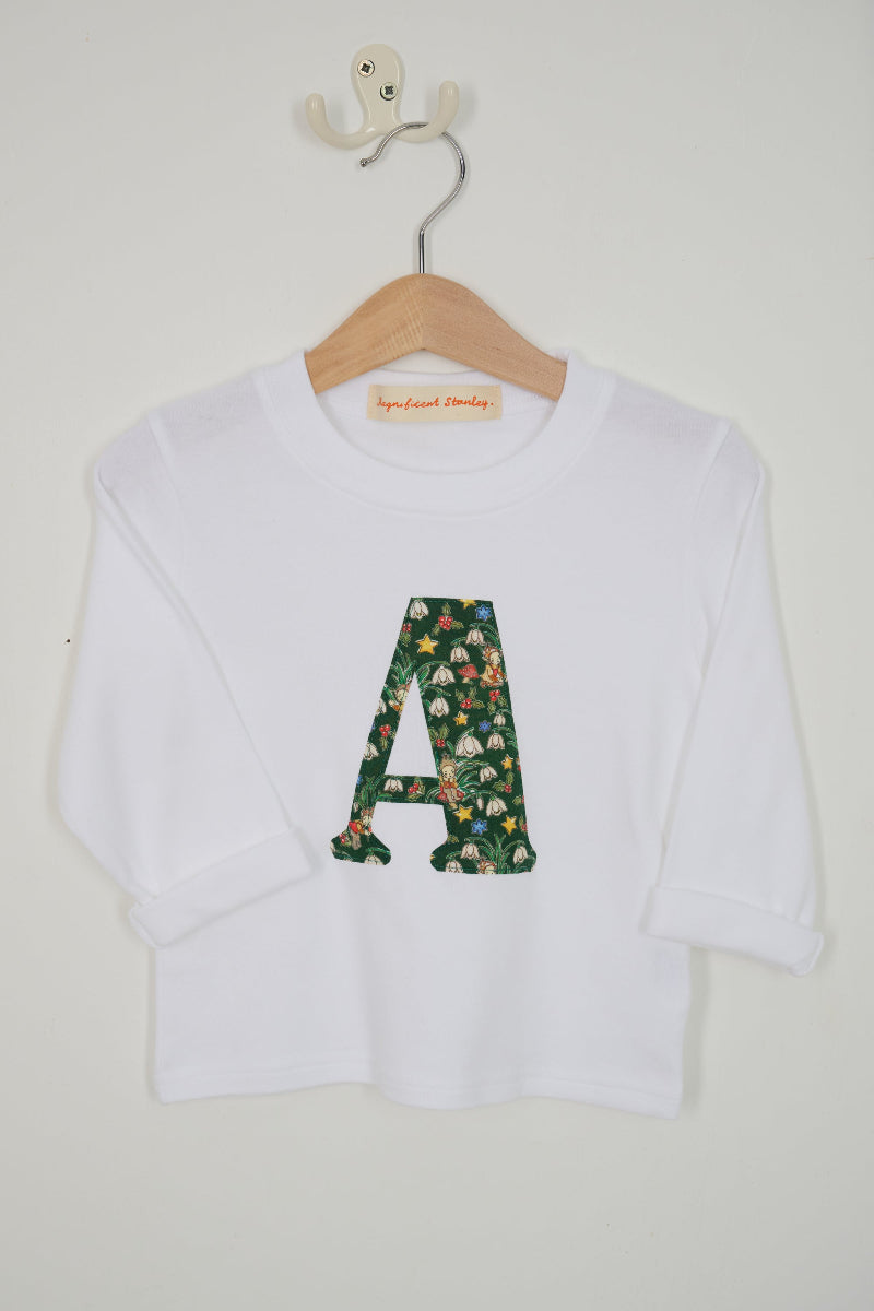 A white long-sleeve top for kids, featuring a large embroidered letter 'A' on the front filled with a green floral Liberty Elves pattern. The shirt is displayed on a wooden hanger with a white wall hook behind it.
