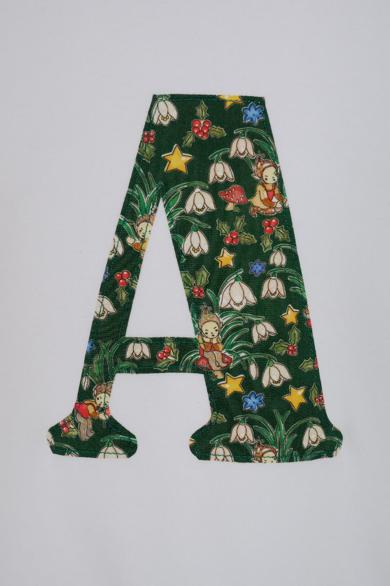 A decorative capital letter A cutout featuring a Liberty Elves pattern with white snowdrops, yellow stars, red berries, and a small elf on a dark green background, shown against a plain light gray backdrop.