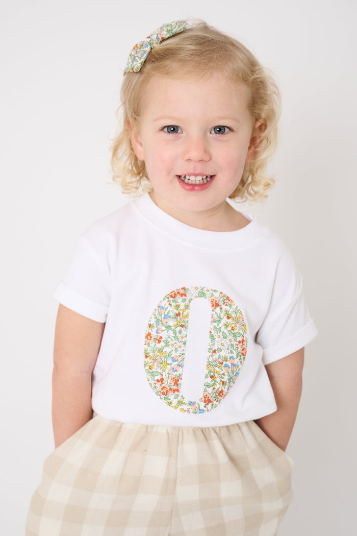 A 2 year old girl wearing a summery personalised white cotton t-shirt with her initial cut from Nazanin floral liberty fabric.