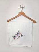 A white baby blanket with a colorful zoo animal collage print, hanging from a wooden hanger on a light gray wall.