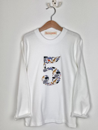  A white kids birthday t-shirt hanging against a white wall. The t-shirt features a number 5 cut from liberty zoo print.