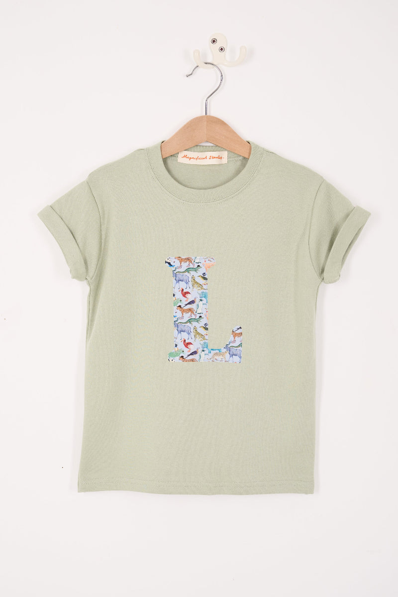 A sage green short-sleeve t-shirt hanging on a wooden hanger against a white wall. The shirt features a large, decorative letter 'L' printed on the chest, filled with a Liberty pattern of  zoo animals.