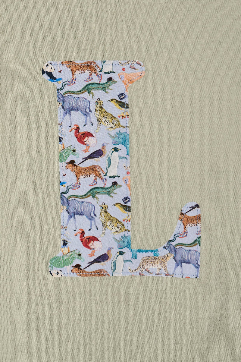 A sage green short-sleeve t-shirt hanging on a wooden hanger against a white wall. The shirt features a large, decorative letter 'L' printed on the chest, filled with a Liberty pattern of  zoo animals.