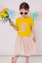 a smiling 3 year old girl wearing a yellow personalised t-shirt with her initial cut from floral liberty print on the front.