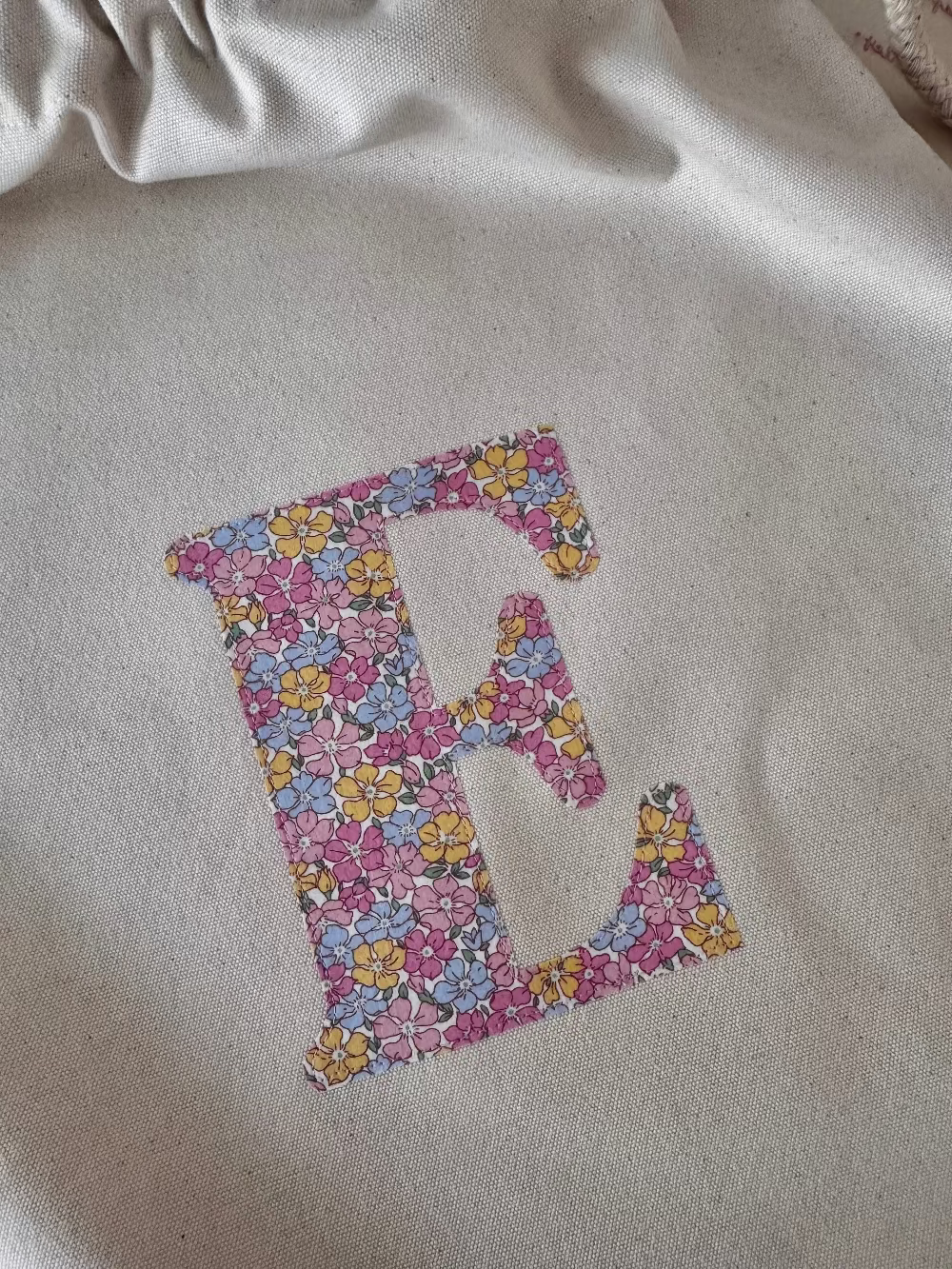 A cream canvas drawstring backpack with a personalised Liberty floral initial sewn on the front